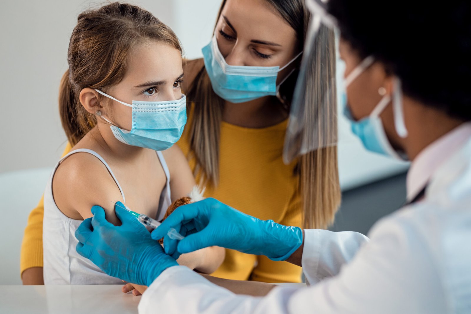 Little girl with protective face mask getting a vaccine while being with her mother at pediatrician's.